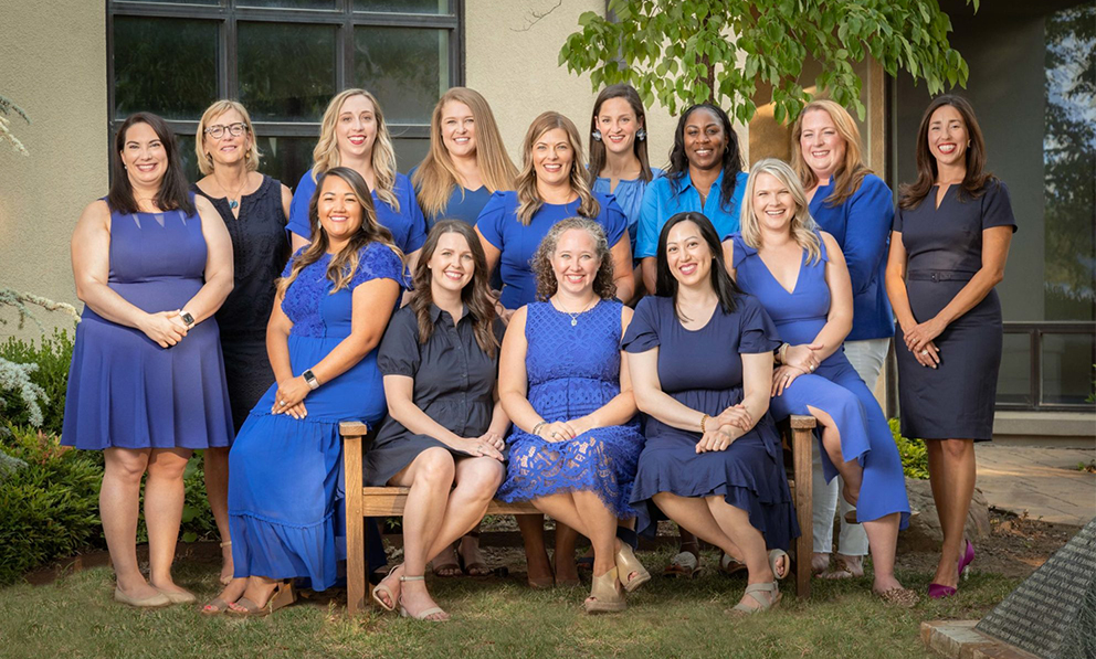 2021-22 Junior League of Tulsa Board of Directors. 14 women, dressed in shades of blue, are arranged in a group formation. The women in front are sitting on a bench.